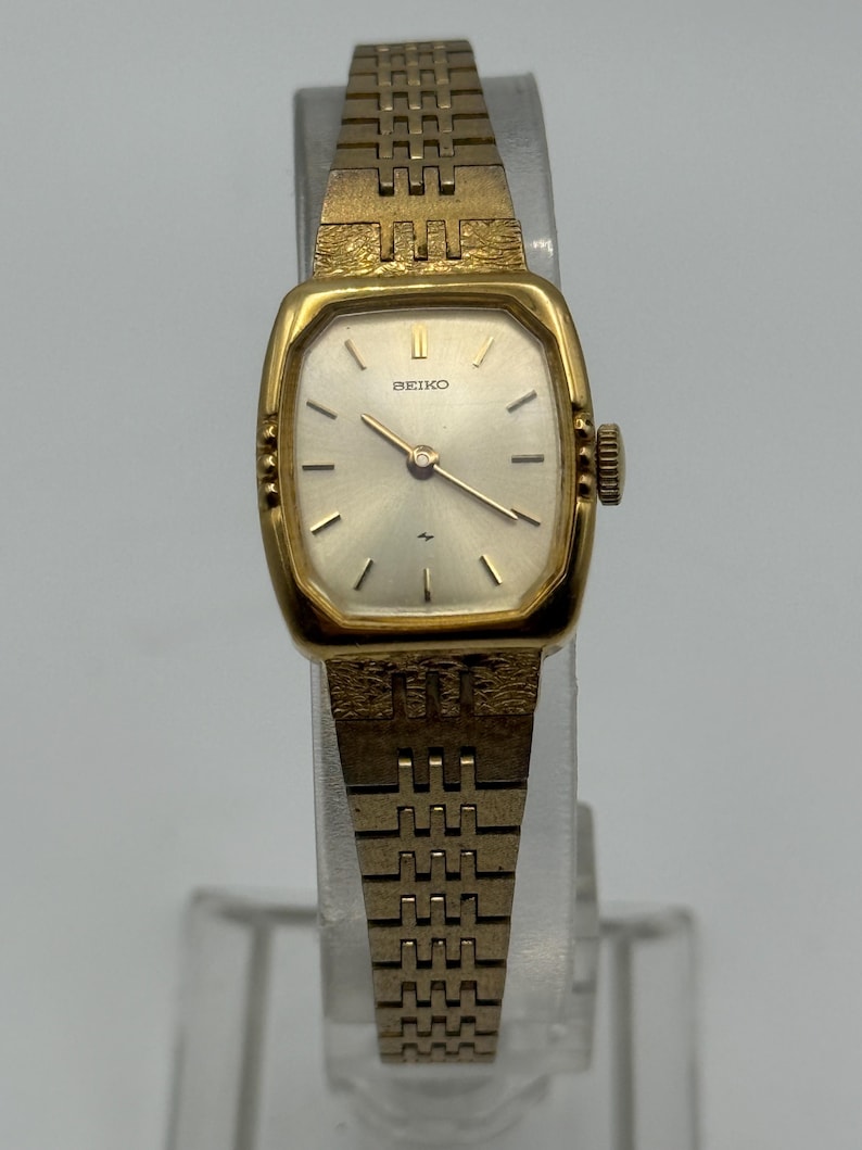 May include: A vintage gold-toned Seiko wristwatch with a rectangular face and a matching link band. The watch face is silver with black hour markers and hands. The watch is displayed on a clear stand.