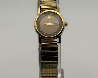 May include: A gold-toned Seiko Quartz wristwatch with a round face and a gold and silver expanding band. The watch face is a dark brown color with gold hands and Roman numeral hour markers. The watch is displayed on a clear stand.