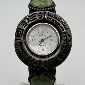May include: A silver-toned BOCCQ Quartz watch with a round face and a decorative bezel. The watch features a mother-of-pearl dial, Roman numeral hour markers, and two oval turquoise-colored stones on the band.