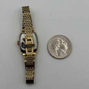 May include: A vintage gold-toned wristwatch with a rectangular face and a linked band. The watch is positioned next to a quarter coin for size comparison. The watch's face is visible, and the band is partially extended.