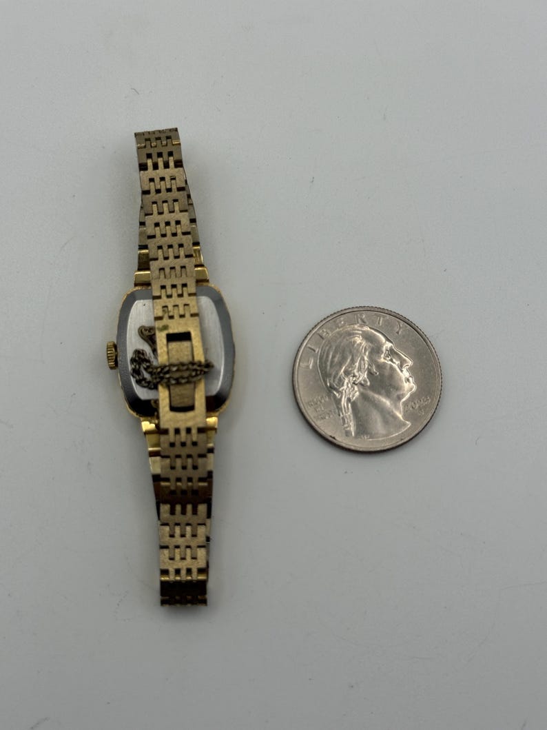 May include: A vintage gold-toned wristwatch with a rectangular face and a linked band. The watch is positioned next to a quarter coin for size comparison. The watch's face is visible, and the band is partially extended.