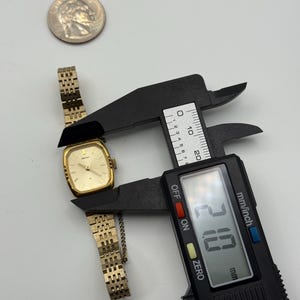 May include: A gold-toned vintage wristwatch with a rectangular face and a linked band. The watch is being measured with a digital caliper, showing a width of 21.0 mm. A silver coin is also present.
