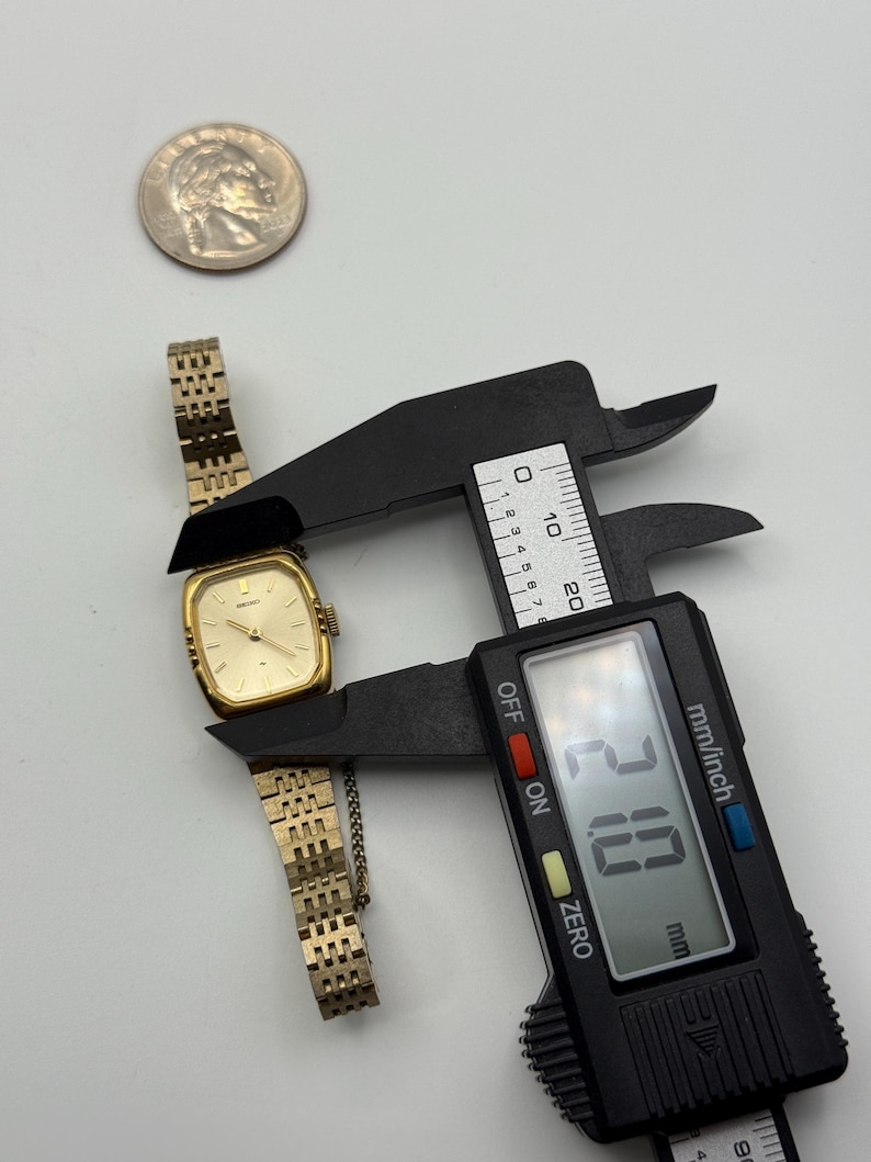 May include: A gold-toned vintage wristwatch with a rectangular face and a linked band. The watch is being measured with a digital caliper, showing a width of 21.0 mm. A silver coin is also present.