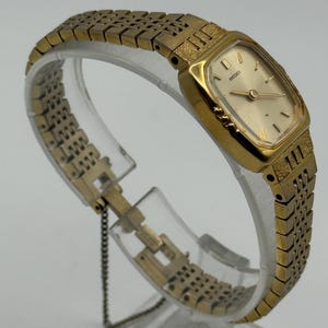 May include: A gold-toned Seiko wristwatch with a rectangular face and a matching gold band. The watch has a light-colored dial with gold hands and markers. The band features a textured design. The watch is displayed on a clear stand.
