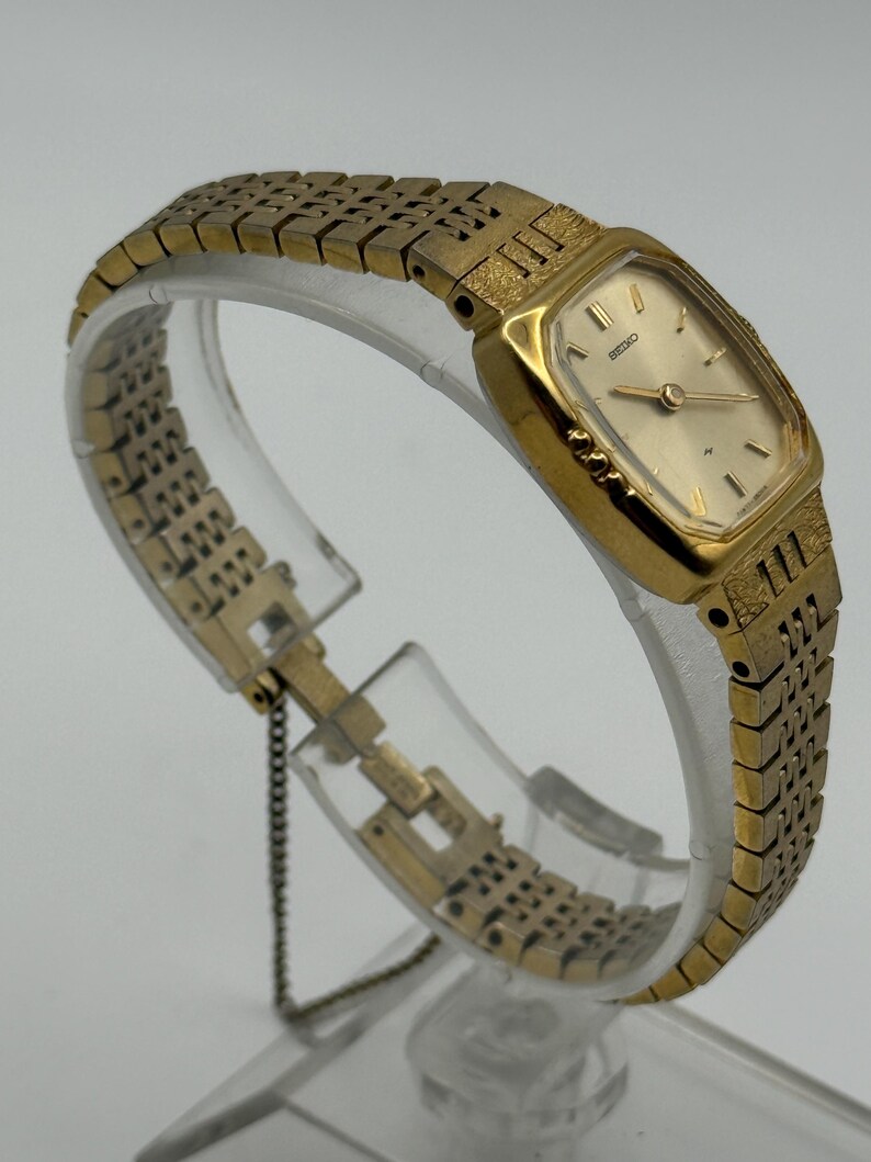 May include: A gold-toned Seiko wristwatch with a rectangular face and a matching gold band. The watch has a light-colored dial with gold hands and markers. The band features a textured design. The watch is displayed on a clear stand.