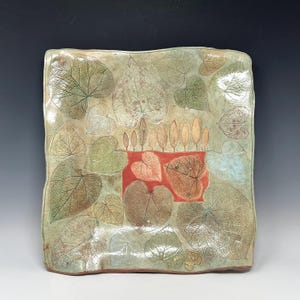 May include: Square ceramic serving dish with a leaf design. The dish features a light green background with various leaf imprints in shades of green, brown, and white. A red square in the center has leaf imprints and a row of stylized trees.