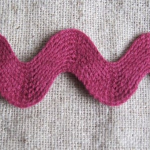 May include: Close-up of a magenta-colored, wavy trim. The trim has a textured, woven appearance and is set against a neutral, burlap-like background. The decorative trim is likely used for sewing or crafting projects.