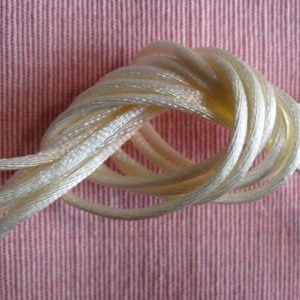 May include: Close-up of a cream-colored rope tied in a decorative knot. The rope is made of multiple strands and has a slightly shiny texture. The background is a pink fabric with vertical stripes.