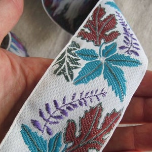 Puede incluir: Cinta blanca con un patrón floral detallado con hojas y ramitas de color turquesa, burdeos y morado. La cinta se sostiene en una mano, mostrando el diseño y la textura del tejido.
