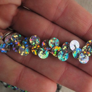 May include: Close-up of a strand of iridescent sequins. The sequins are round, with a shiny, reflective surface that shimmers with multiple colors. The sequins are sewn onto a thin, white thread, creating a decorative trim.