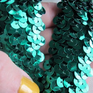 以下が含まれることがあります： エメラルドグリーンのスパンコール2本のクローズアップ。各スパンコールは丸く光沢があり、中央に小さな穴があります。スパンコールは列に配置され、テクスチャード加工されたきらめく効果を生み出しています。背景は柔らかくニュートラルな色です。