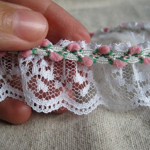 May include: White lace trim with a scalloped edge, accented with a band of pink and green floral embellishments. The delicate trim is held by a hand, showcasing the intricate details of the lace and floral design.