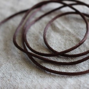 May include: Close-up of a length of dark brown cord, coiled and resting on a neutral-colored fabric. The cord appears to be made of a woven material, with a slightly textured surface. The lighting is soft, highlighting the cord's color and texture.