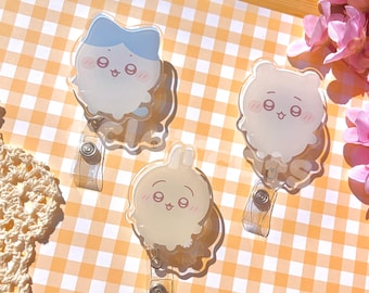 May include: Three acrylic badge reels featuring cartoon characters with expressive faces. One character has blue hair, while the others are cream-colored with rounded ears. Each reel includes a clear plastic strap and a metal button mechanism.