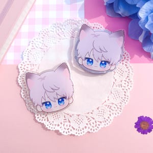 Peut inclure: Deux breloques en acrylique transparent repr&eacute;sentant un gar&ccedil;on-chat de dessin anim&eacute; avec des cheveux violets clairs, des yeux bleus et des oreilles de chat. Les breloques sont pos&eacute;es sur une nappe en dentelle blanche, avec un fond &agrave; carreaux roses et blancs.