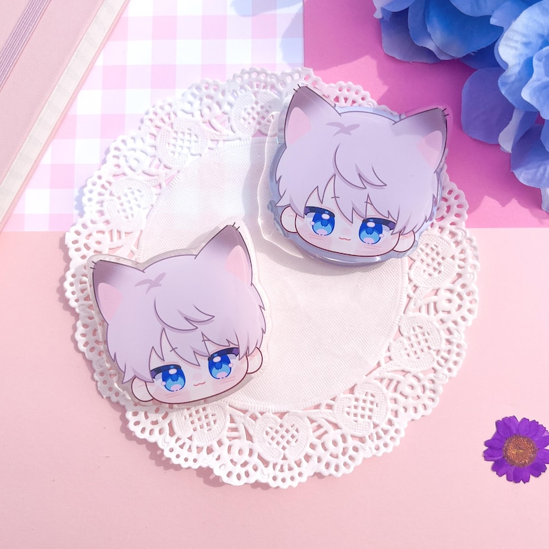 Peut inclure: Deux breloques en acrylique transparent repr&eacute;sentant un gar&ccedil;on-chat de dessin anim&eacute; avec des cheveux violets clairs, des yeux bleus et des oreilles de chat. Les breloques sont pos&eacute;es sur une nappe en dentelle blanche, avec un fond &agrave; carreaux roses et blancs.