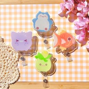 Könnte beinhalten: Vier Ausweishalter aus Acryl mit Zeichentrickfiguren: ein hellblauer Relaxo, ein hellviolettes Gengar, ein leuchtend orangefarbener Goldfisch und ein hellgrünes pflanzenartiges Wesen, jeweils mit einem transparenten Kunststoffband.