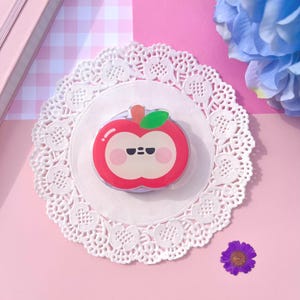 May include: A red apple-shaped phone grip with a cute face, green leaf, and orange stem. The grip is on a white doily, set against a pink background with a small purple flower and a blue flower.