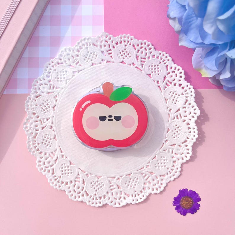 May include: A red apple-shaped phone grip with a cute face, green leaf, and orange stem. The grip is on a white doily, set against a pink background with a small purple flower and a blue flower.