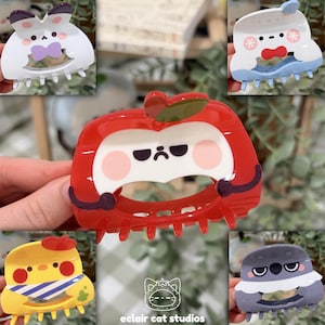 May include: A collection of colorful, cartoon-style hair claw clips. The clips feature various designs, including a white bunny with a purple bow, a red apple with a face, and a yellow chick with a blue and white scarf. The clips are made of plastic.