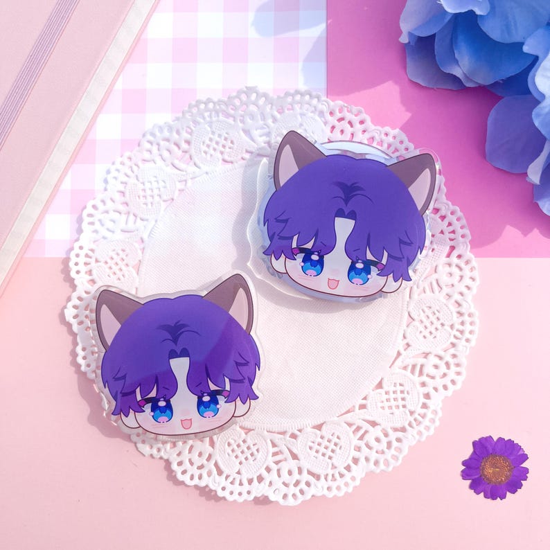 Peut inclure: Deux breloques en acrylique transparent repr&eacute;sentant un personnage de dessin anim&eacute; aux cheveux violets, aux yeux bleus et aux oreilles de chat. Les breloques sont pos&eacute;es sur une nappe en dentelle blanche, sur fond rose avec une petite fleur violette et des accents floraux bleus.