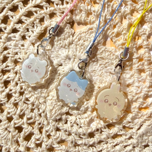 May include: Three acrylic charms featuring cartoon characters with blushing cheeks and open mouths. Each charm hangs from a metal lobster clasp attached to a thin nylon cord in pink, blue, and yellow. The charms have a glossy, transparent finish.