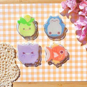 May include: Four circular acrylic phone grips featuring cartoon character designs. The top left is a light green creature with leaves, the top right is a blue and white creature, the bottom left is a light purple creature, and the bottom right is an orange fish.