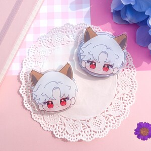 Peut inclure: Deux breloques en acrylique transparent repr&eacute;sentant un personnage de dessin anim&eacute; aux cheveux blancs, aux oreilles marron et aux yeux rouges. Les breloques sont pos&eacute;es sur une nappe en dentelle blanche, sur fond rose &agrave; carreaux.