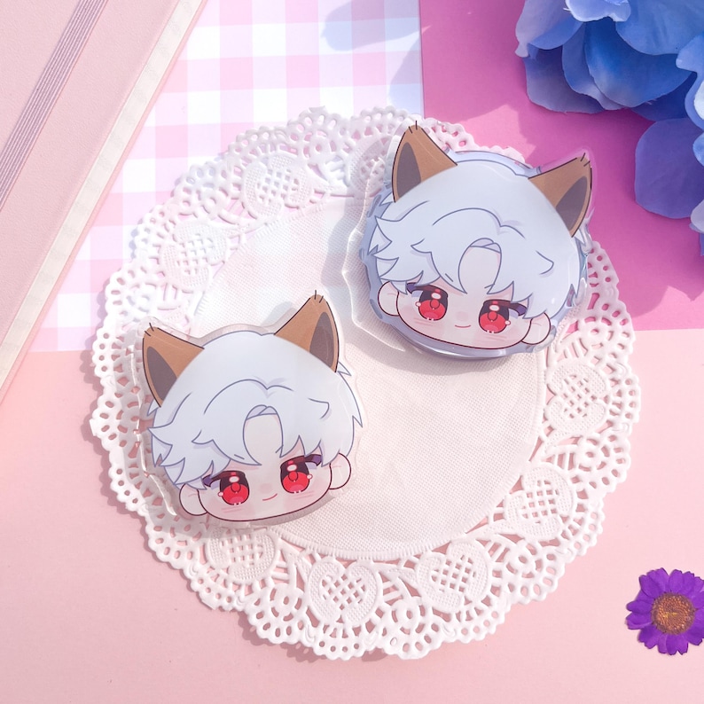 Peut inclure: Deux breloques en acrylique transparent repr&eacute;sentant un personnage de dessin anim&eacute; aux cheveux blancs, aux oreilles marron et aux yeux rouges. Les breloques sont pos&eacute;es sur une nappe en dentelle blanche, sur fond rose &agrave; carreaux.