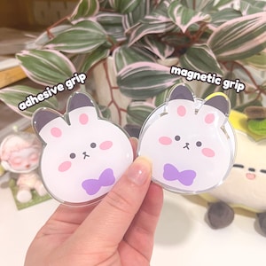 May include: Two clear, bunny-shaped phone grips. One is labelled "adhesive grip" and the other "magnetic grip." The bunnies have white bodies, pink cheeks, black eyes, and purple bow ties. The grips are held in a hand.