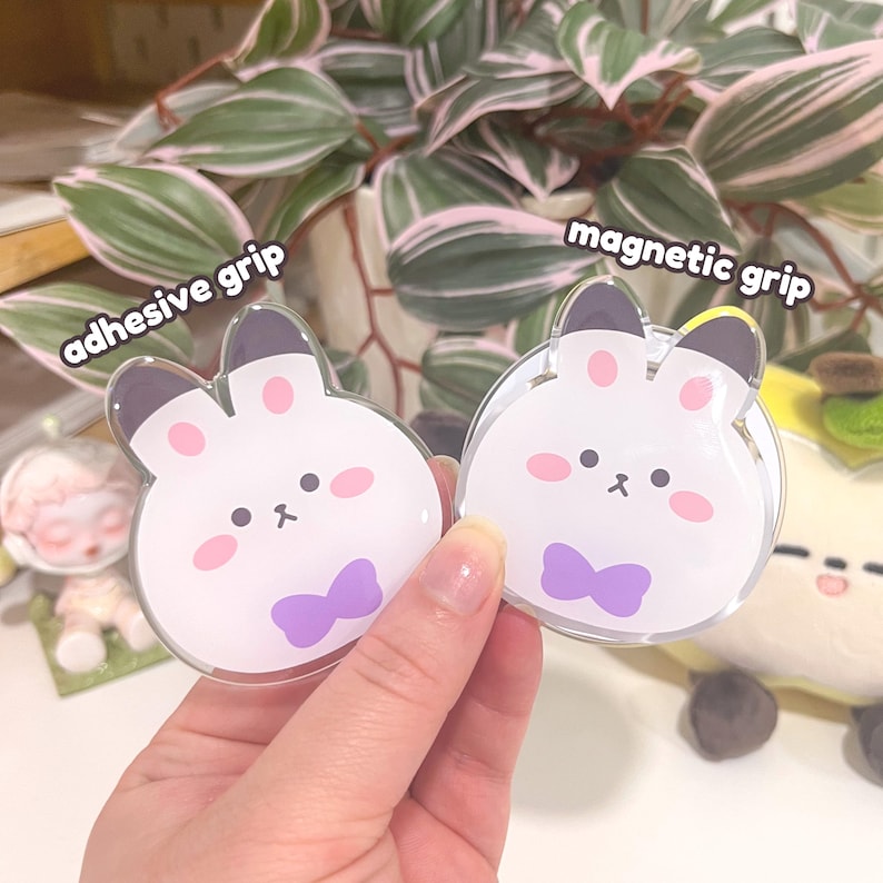 May include: Two clear, bunny-shaped phone grips. One is labelled "adhesive grip" and the other "magnetic grip." The bunnies have white bodies, pink cheeks, black eyes, and purple bow ties. The grips are held in a hand.