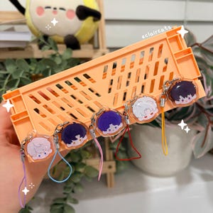 May include: A collection of anime-style character charms with various hair colors, each attached to a keychain and colorful lanyard. The charms are displayed in an orange plastic crate. The text "eclaircats" is visible.