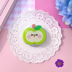 May include: A green apple-shaped decorative item with a cute face, a red stem, and a green leaf. The item is placed on a white doily with a pink background. The apple has a light green body with a white centre.