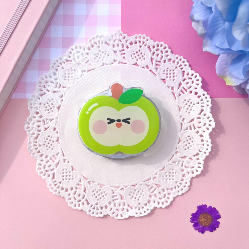 May include: A green apple-shaped decorative item with a cute face, a red stem, and a green leaf. The item is placed on a white doily with a pink background. The apple has a light green body with a white centre.