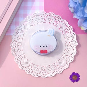 May include: A white, cartoon-style phone grip featuring a bunny character with a popsicle on its head and a red bow tie. The grip is on a white lace doily, with a pink and white checkered background.