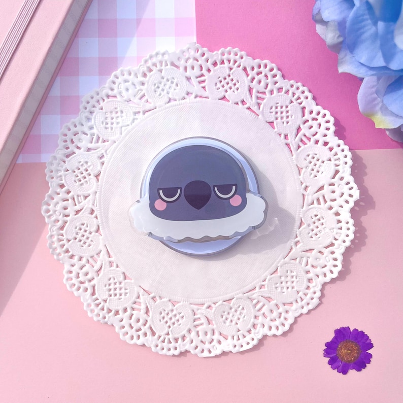 May include: A circular sticker with a cartoon bird design, in shades of grey and white, with a grumpy expression. The sticker is placed on a white doily, with a pink background and a small purple flower.