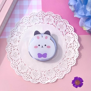 May include: A white bunny phone grip with a purple bow tie, placed on a white lace doily. The bunny has black ears and pink cheeks. The background is pink and includes a purple flower and a blue flower.