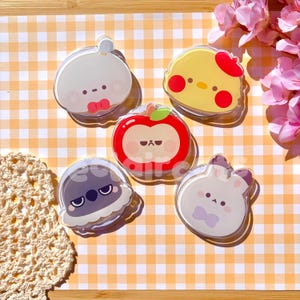 May include: Five circular acrylic phone grips featuring cartoon characters: a white creature with a red bow, a yellow chick with a red hat, a red apple with a face, a gray bird with a grumpy expression, and a white rabbit with a purple bow.