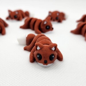 Könnte beinhalten: Eine Gruppe von sieben kleinen, braunen, 3D-gedruckten Fuchsfiguren mit schwarzen Augen und weißen Akzenten. Die Füchse sind in verschiedenen Positionen platziert, einige liegen, andere stehen.