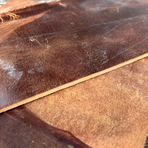 May include: Close-up of two pieces of brown leather. The leather has a distressed appearance with visible grain and variations in colour, ranging from light tan to dark brown. The edges are cleanly cut, and the surface shows signs of wear.