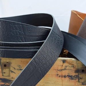 May include: Close-up of black leather strips, one with a textured pattern, coiled on a wooden crate. Additional leather pieces in brown and black are visible in the background. The leather appears to be for crafting or design.