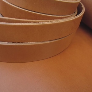 May include: Close-up of coiled, light brown leather strips, likely for crafting or leatherwork. The strips are stacked, revealing their thickness and texture. The background shows a smooth, matching leather surface.