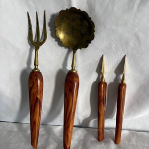 Peut inclure: Un ensemble d'ustensiles de service anciens, comprenant une fourchette, une cuillère et deux couteaux. Les ustensiles ont des têtes en laiton et des manches avec une finition marbrée de couleur ambre. La cuillère a un bol festonné décoratif.