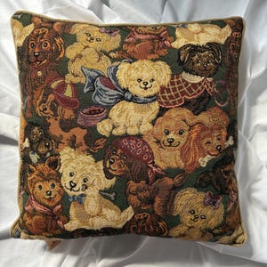 Peut inclure: Coussin carré avec un motif de tapisserie représentant diverses races de chiens dans des tons de marron, beige et crème sur un fond vert foncé. Le coussin a une bordure dorée.