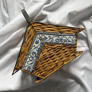 May include: Woven wicker basket with a decorative blue and white tile border. The basket has a metal handle and an angular shape. The tile features a floral design. The basket is likely for decorative or storage use.