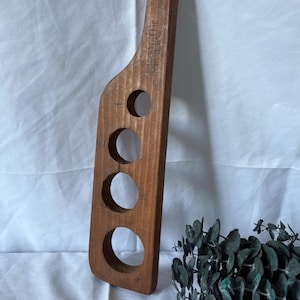 Pode incluir: Medidor de espaguete de madeira com três orifícios circulares e a palavra "spaghettii" gravada na lateral. A ferramenta é feita de madeira castanha clara e tem um orifício na parte superior para pendurar. Uma planta verde decorativa está no fundo.