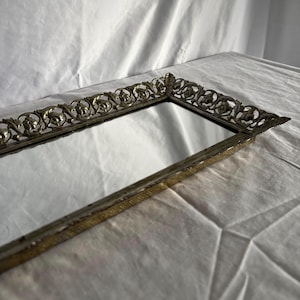 Peut inclure: Miroir de coiffeuse rectangulaire antique avec un cadre en métal doré orné de motifs floraux. La surface réfléchissante du miroir est entourée du cadre décoratif, ajoutant une touche d'élégance vintage. Le design complexe du cadre est visible.