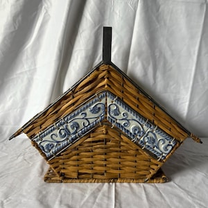 May include: A woven wicker basket with a roof-like design. The roof features decorative blue and white tiles. A black metal handle extends from the top. The basket has a rectangular base and is set against a white background.