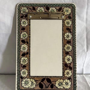 Puede incluir: Un portapapeles rectangular con un diseño floral. El borde es marrón con un patrón repetido de flores blancas y verdes. El clip es plateado y el interior es de color crema.
