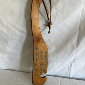 May include: Wooden oven rack puller with a curved handle and a leather loop. The puller has the words "OVEN RACK PULLER" printed in blue, along with blue heart designs. The puller is approximately 30 cm long.